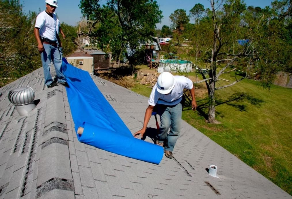 Roofers unrolling blue tarp on gray shingle roof