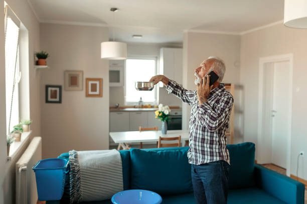 Angry Bearded Senior Man Talking on Smartphone Near Plastic Wash Bowl at Home in the Living Room Because of Roof Leaking